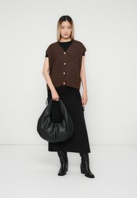 Eine Frau trägt eine braune ärmellose Knopf-Weste über einem schwarzen Kleid, hält eine große schwarze Lederhandtasche und trägt schwarze Stiefeletten mit Absatz.