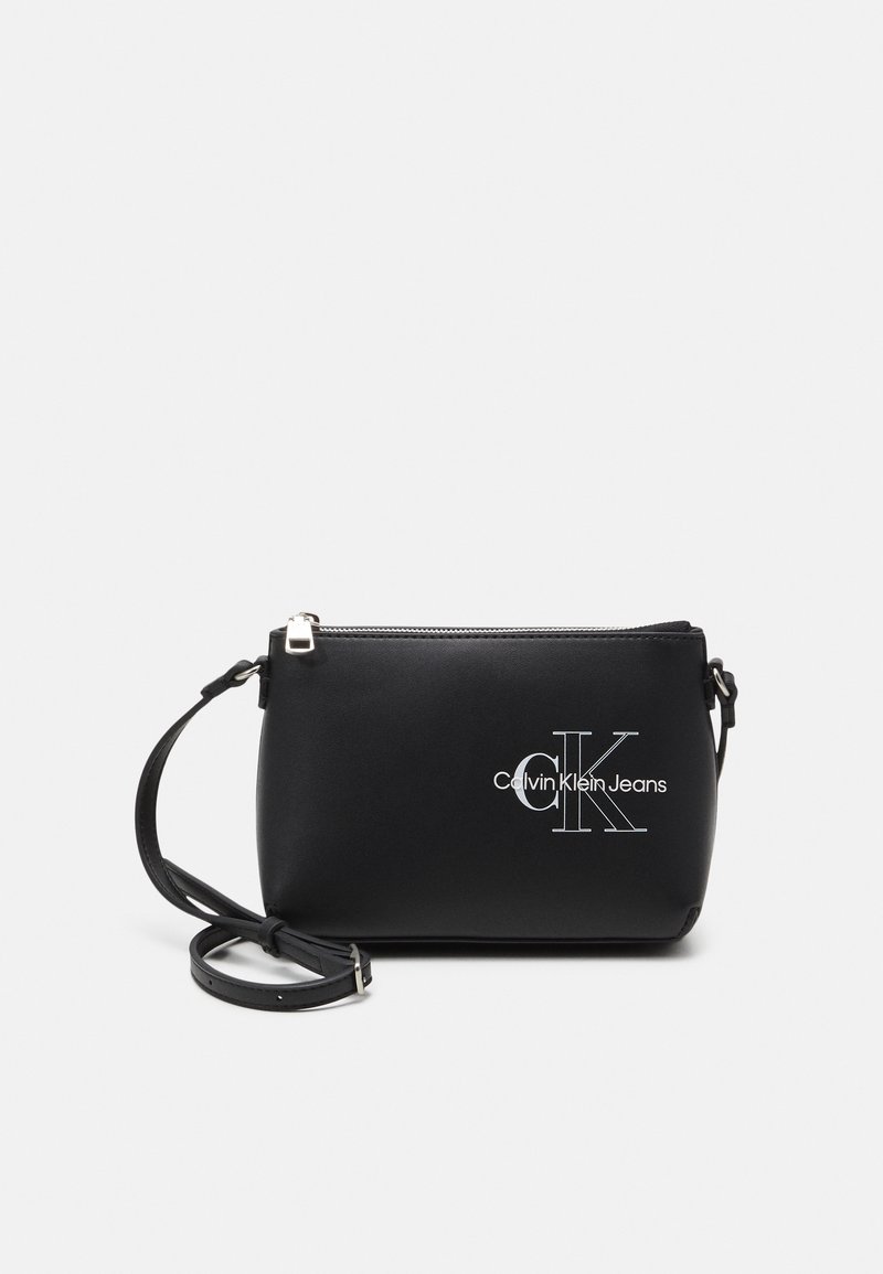 Musta synteettinen olkalaukku, jossa on hopeanvärinen vetoketjusuljenta, valkoinen "Calvin Klein Jeans" -logo ja säädettävä hihna.