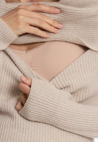 Hellbeige gestrickter Pullover mit gerippter Textur und weiten Bündchen, die eine hellrosa Basisschicht darunter sichtbar machen. Nahaufnahme von Stoff und Händen.