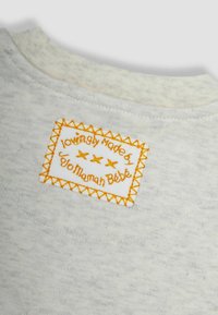 Ein weißer Stoff mit einer weichen Textur zeigt ein sichtbares Etikett mit orangefarbener Stickerei, auf dem steht "liebevoll hergestellt von Jojo Maman Bébé", umrandet von Zickzackstichen.