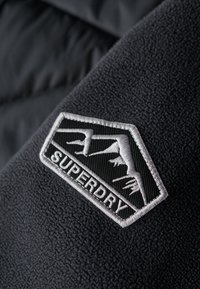 Material de forro polar gris oscuro con una superficie texturizada, que presenta un parche de logotipo bordado en blanco y negro de la marca Superdry.