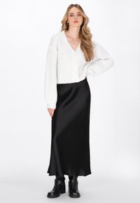 Cardigan tricotat alb, scurt, cu decolteu în V și nasturi frontali, asociat cu o fustă lungă din satin negru și cizme negre până la glezne.