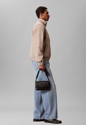 Jonge man in een beige jas en jeans met wijde pijpen die een kleine zwarte handtas vasthoudt, staand tegen een grijze achtergrond.