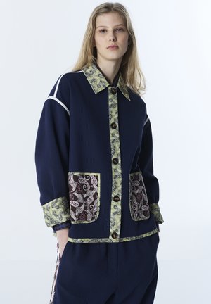 Młoda kobieta ubrana w granatowy strój z kołnierzem, mankietami, listwą z przodu i obramowaniem kieszeni w wzór paisley, stojąca na tle gładkiej białej ściany.