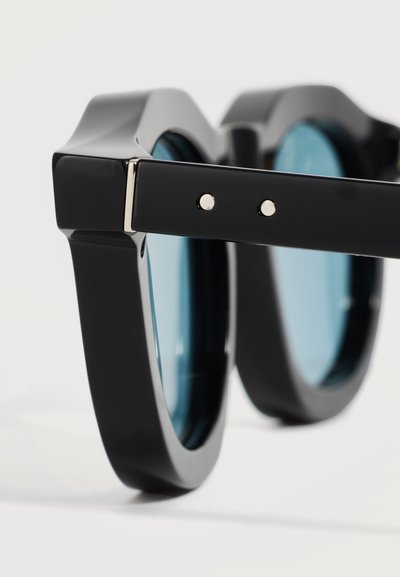 Gros plan de lunettes de soleil noires brillantes à monture épaisse avec des verres teintés bleus et deux petits points métalliques sur la branche.