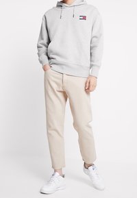 Felpa grigia con tasca frontale e patch del logo; pantaloni beige a sigaretta; sneakers bianche con dettagli in rete. Tessuto morbido, design informale.