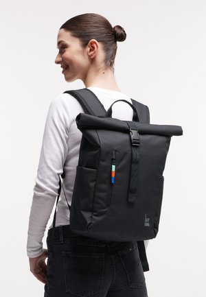 Vrouw met donker haar in een knot, gekleed in een wit shirt met lange mouwen en zwarte spijkerbroek, met een zwarte roll-top rugzak met kleurrijke rits.