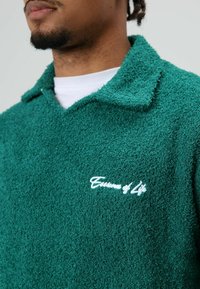 Groene getextureerde pullover met kraag gemaakt van zachte stof, met een geborduurd wit logo op de linkerborst, gedragen over een wit overhemd.