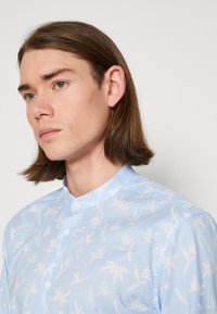 Camicia azzurro chiaro con motivi a foglia bianchi, colletto alla mandarina abbottonato e una superficie liscia. Presenta una vestibilità rilassata e un tessuto morbido.