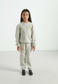 Ragazzina in piedi, con indosso un completo coordinato di felpa e pantaloni della tuta grigi chiari e scarpe slip-on nere, su uno sfondo bianco uniforme.