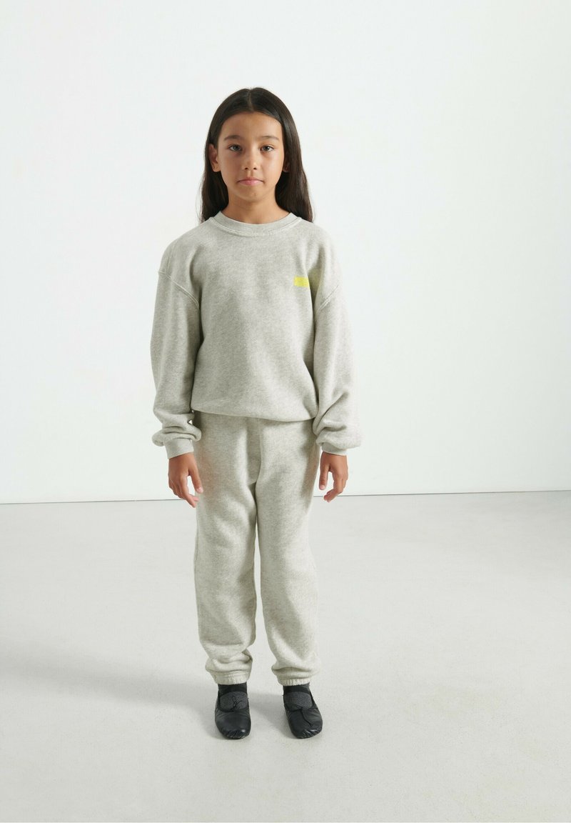 Ragazzina in piedi, con indosso un completo coordinato di felpa e pantaloni della tuta grigi chiari e scarpe slip-on nere, su uno sfondo bianco uniforme.