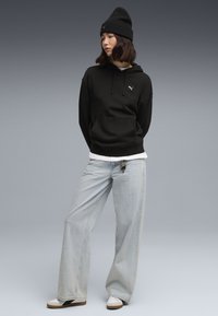 Čierna mikina s kapucňou a predným vreckom a logom, v kombinácii s svetlomodrými širokými džínsami a čiernou čiapkou. Biely sneakers dotvárajú outfit.