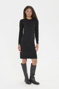 Göndör hajú nő fekete, testhezálló, hosszú ujjú ruhát és térdig érő, fekete bőr csizmát viselve áll fehér háttér előtt.