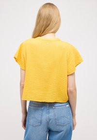 Gul tekstureret cropped top med korte ærmer, der har en lille detalje bagpå, parret med lyseblå højtaljede jeans.