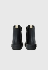 Bottines noires en cuir avec une finition lisse, un accent doré sur la languette, et des semelles en caoutchouc texturé. Logo de la marque sur le talon.