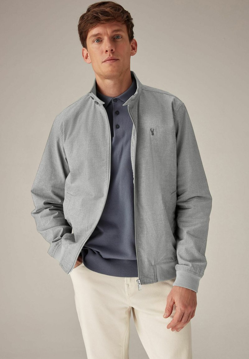 Chaqueta gris con cremallera y cuello alzado, de tela texturizada, con un pequeño logo en el pecho, llevada sobre una camiseta polo azul marino y pantalones claros.