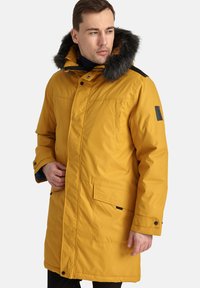 Parka isolata gialla con cappuccio in pelliccia sintetica rimovibile, chiusure a scatto, tasche laterali e dettagli neri a contrasto sulle spalle.