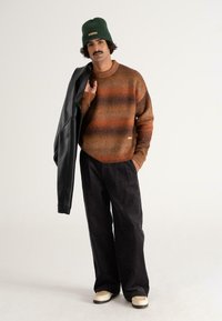 Strickpullover mit braunen und orangefarbenen Streifen, schwarze plissierte Hose und eine grüne Mütze. Trägt eine schwarze Lederjacke über einer Schulter.