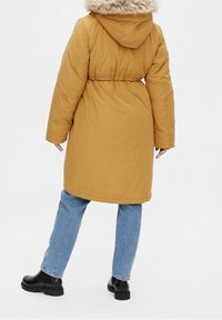 Hořčičková parka s kapucí lemovanou kožešinou, staženým pasem a dlouhými rukávy. Nositá přes modré džíny a černé kotníkové boty.