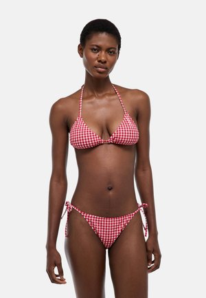 Femme portant un bikini à carreaux rouges et blancs avec un haut licou et un bas noué sur les côtés, debout devant un fond clair uni.