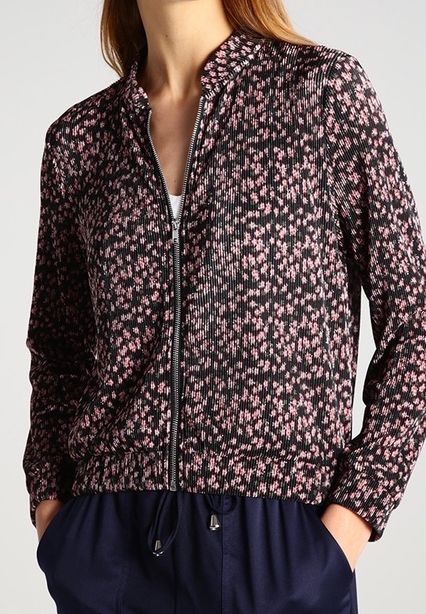 Femme portant une veste noire zippée à motif floral rose et un pantalon bleu marine à cordon, les mains dans les poches, debout devant un fond uni.
