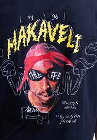 Svart t-shirt med ett grafiskt tryck av en person med solglasögon och en röd bandana. Texten inkluderar "MAKAVELI" och "THUG LIFE" i gult.