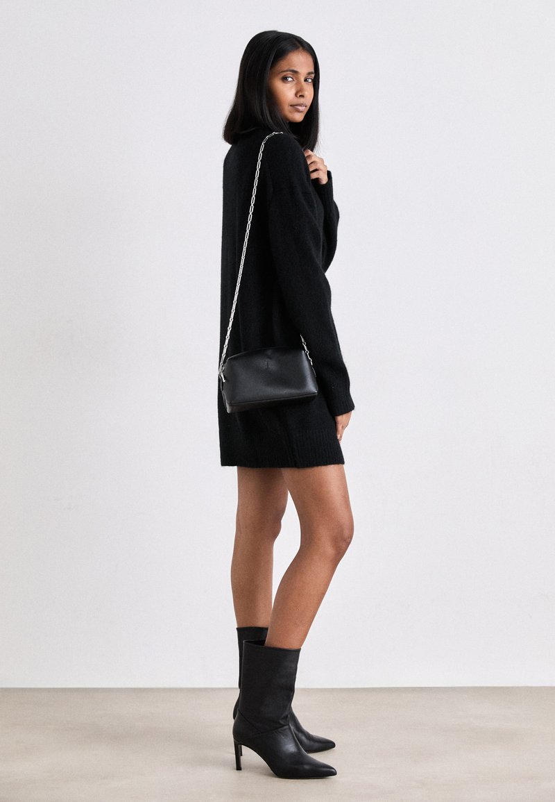 Black leather crossbody bag with silver chain strap, rectangular shape, smooth texture. Model wears black knit dress and knee-high heeled boots.