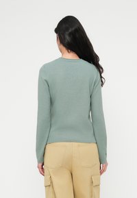 Πουλόβερ μακρυμάνικο σε μέντα πράσινο χρώμα με στρογγυλή λαιμόκοψη, απαλό υλικό και εφαρμοστή γραμμή, συνδυασμένο με μπεζ παντελόνι καμπαρντίνα με πίσω τσέπες.