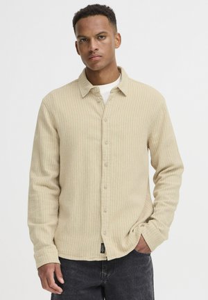 Beige stribet skjorte med knaplægning, krave og lange ærmer, der har et struktureret stof og subtile farvevariationer. Sorte jeans komplementerer outfittet.
