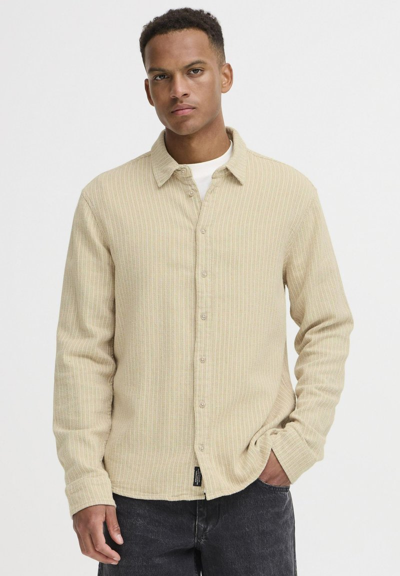 Beige stribet skjorte med knaplægning, krave og lange ærmer, der har et struktureret stof og subtile farvevariationer. Sorte jeans komplementerer outfittet.