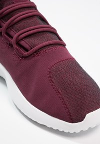 Vínová sportovní bota s texturovaným pleteným svrškem, elastickými šňůrkami a kontrastní bílou podrážkou. Má jemné švy a hladký povrch.