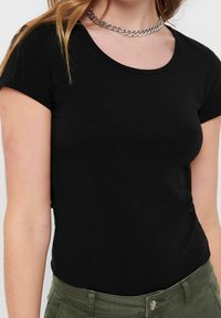 Svart kortärmad t-shirt med rundad halsringning, tillverkad av mjukt tyg. Matchas med gröna byxor, vilket framhäver en avslappnad look. Kedjehalsband som accessoar.