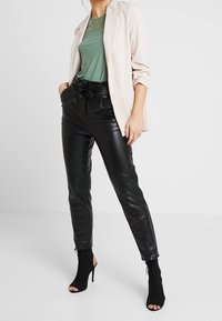 Blazer beige clair, haut vert avec détail en dentelle, pantalon en cuir noir ajusté, et bottines noires ouvertes à l'avant avec fermetures éclair.