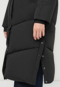 Nærbillede af en persons hånd, der hviler på en sort pufferjakke med trykknapper og slidser i siden, hvor der ses blå denim under.