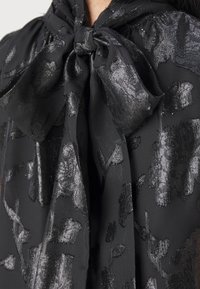 Tissu noir transparent avec un motif floral métallique noué en un grand nœud au niveau de l'encolure.