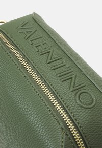 Πράσινη δερμάτινη τσάντα χειρός με ανάγλυφο λογότυπο "Valentino", χρυσό κλείσιμο με φερμουάρ και υφή επιφάνειας. Κοντινή λήψη των λεπτομερειών σχεδίασης.