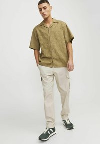 Camisa de manga corta color oliva con un patrón geométrico, combinada con pantalones cargo en un tono beige claro y zapatillas de deporte verde oscuro con acentos blancos.