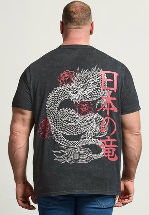 Mand iført sort T-shirt med stort hvidt drage- og rødt blomsterprint samt røde japanske tegn på ryggen, stående mod en ensfarvet baggrund.