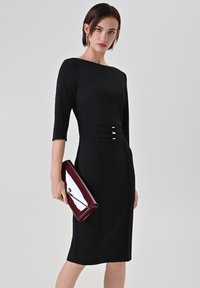 Robe noire ajustée avec manches trois-quarts, dotée d'une ceinture soulignée par deux sangles horizontales et des accessoires dorés. Une pochette bordeaux brillante complète le look.
