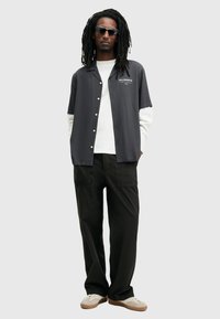 Zwarte shirt met korte mouwen en witte mouwen, met knoopsluiting en logo. Zwarte broek, lichtgekleurde schoenen en een zonnebril.