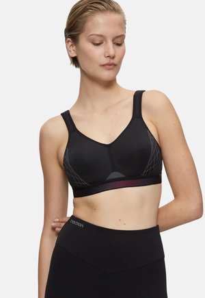 Femme aux cheveux courts portant un soutien-gorge de sport noir avec des côtés à motifs et des leggings noirs taille haute, regardant vers le bas, la main derrière le dos.