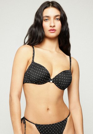 Femme aux longs cheveux foncés portant un haut et un bas de bikini noir à pois sur un fond clair uni.