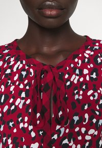 Punainen paita, jossa on sekoitettu leopardikuvio mustilla, valkoisilla ja vaaleansinisillä aksenteilla. Rakenteellinen kangas ja nauhatasku.