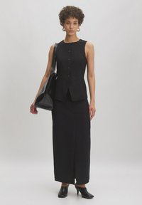 Gilet sans manches noir avec des détails de boutons associé à une longue jupe noire, complété par un cabas noir brillant et des mules à talons noirs.