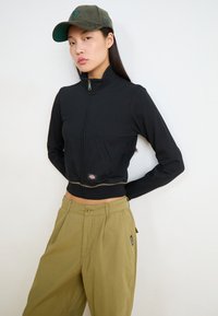 Μαύρο κοντό μπουφάν με φερμουάρ και ψηλό γιακά, με μπροστινές τσέπες και μικρή παθητική λογότυπο; συνδυασμένο με φαρδιές χακί παντελόνες.