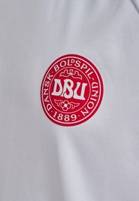 Vit t-shirt med en röd cirkulär logotyp som har "DBU," texten "DANSK BOLDSPIL-UNION," och "1889" i fet stil. Slät yta.