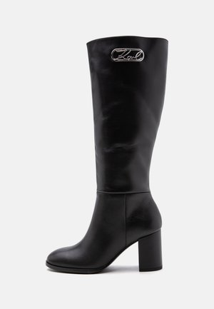 Botte noire en cuir allant jusqu'au genou avec talon bloc et plaque logo en métal argenté près du haut sur le côté extérieur.