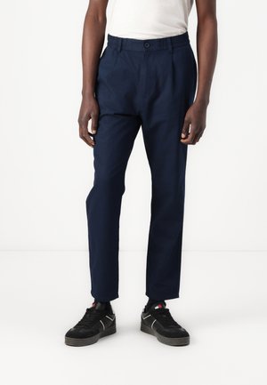Man draagt marineblauwe rechte broek, wit topje en zwarte sneakers, staat tegen een witte achtergrond.