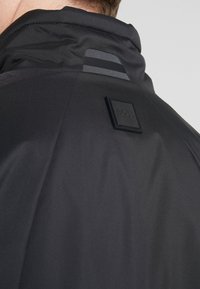 Černá voděodolná bunda s vysokým límcem, která má gumovou logo nášivku a jemné pruhované akcenty na zadní straně. Hladká textura.