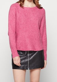 Pull en tricot rose vif à coupe ample, col rond et texture côtelée, associé à une mini jupe en cuir noire avec une fermeture éclair à l'avant.